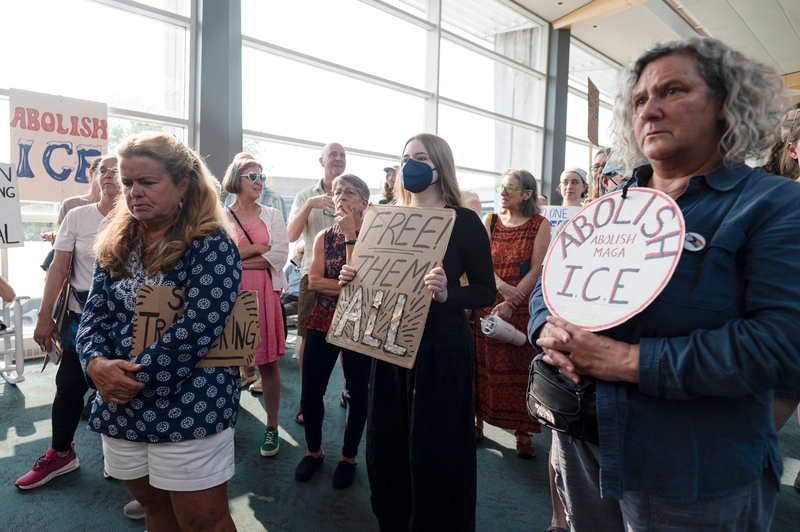 A group of people stand indoors holding protest signs with messages such as "Free Them All" and "Abolish ICE." Some people appear somber, and one wears a face mask.