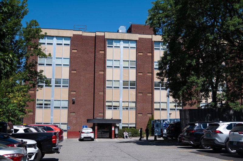 A five-story brick building with vertical beige panels, surrounded by parked cars and trees, under a clear blue sky.