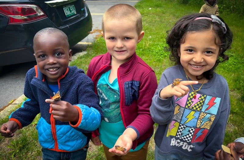 Three young children stand outdoors on grass, smiling and holding butterflies on their hands. A black car and a fourth child are visible in the background.