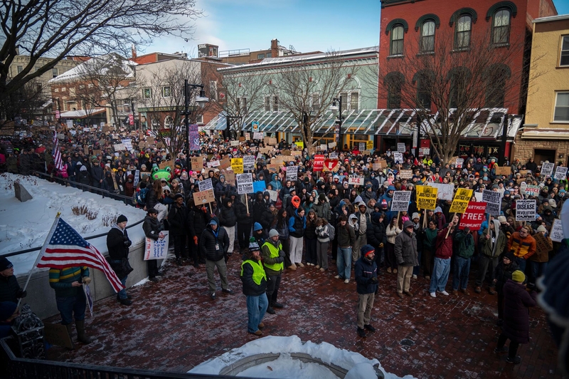 A large crowd gathers outdoors in winter, many holding protest signs and an American flag, with snow on the ground and buildings in the background.