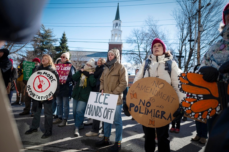A group of people protest outdoors holding signs with messages such as "ICE HANDS OFF," "NO MORE DEPORTATIONS," and "NO AMENDMENT NO MÁS POLIMIGRA." A church is visible in the background.