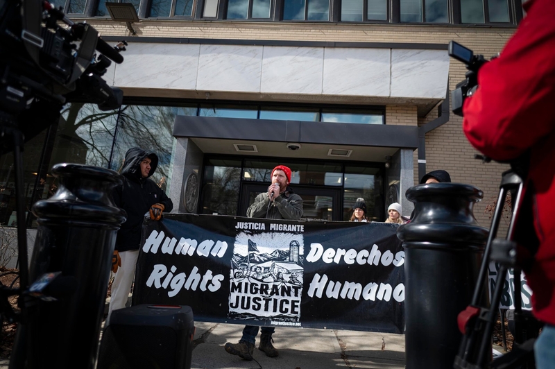 A group of people stand outside a building holding a "Migrant Justice" banner reading "Human Rights" and "Derechos Humanos" during a public demonstration.