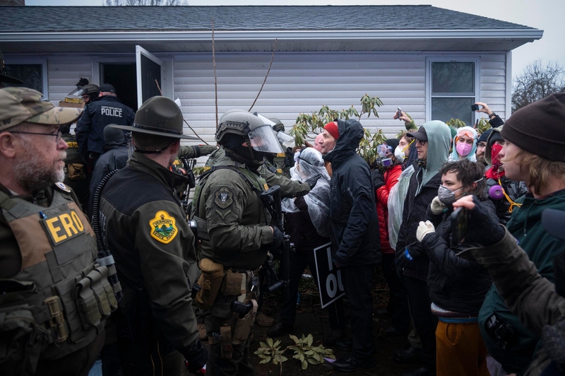 Police officers in tactical gear face a crowd of protesters outside a house; some protesters hold signs and take photos while officers block the entrance.