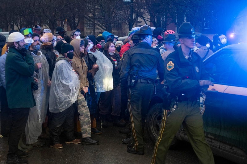 A group of people wearing masks and ponchos face police officers near a patrol car with flashing lights during a protest on a city street.