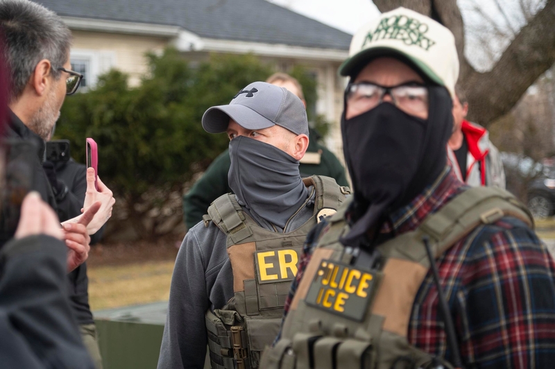 Two people wearing tactical vests labeled "POLICE ICE" and "ERS" stand outdoors, one wearing a mask and hat, as another person gestures with a smartphone nearby.
