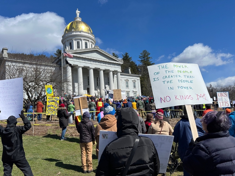 A crowd gathers outside a domed government building holding protest signs, including messages like "NO KINGS" and "The power of the people is greater than the people in power.