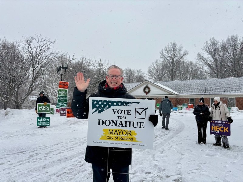 A person stands outside in the snow, smiling and waving, holding a "Vote Tom Donahue Mayor City of Rutland" sign. Other people and campaign signs are visible in the background.
