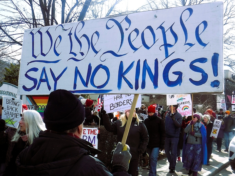 A group of protesters march outdoors holding signs, including a large one that reads "We the People Say No Kings!" in bold lettering.
