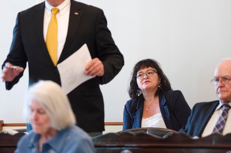 A woman sits in a courtroom, looking forward, while a man in a suit stands holding papers. Two other people are seated nearby, one in the foreground and one to the right.