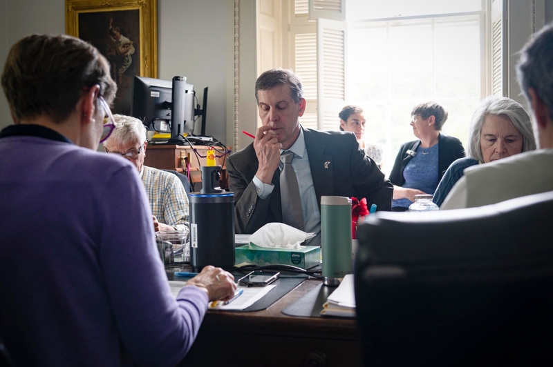 A group of people sitting around a table in a well-lit office, with papers, cups, and a computer visible; one man in a suit is holding a pencil to his lips in thought.