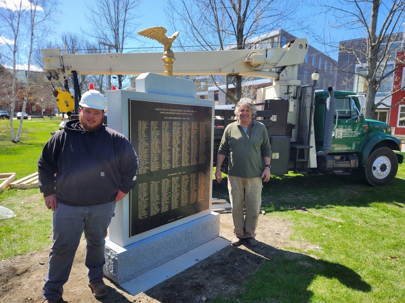 Two men stand next to a newly installed monument with a gold eagle on top; a utility truck and equipment are in the background on a sunny day.