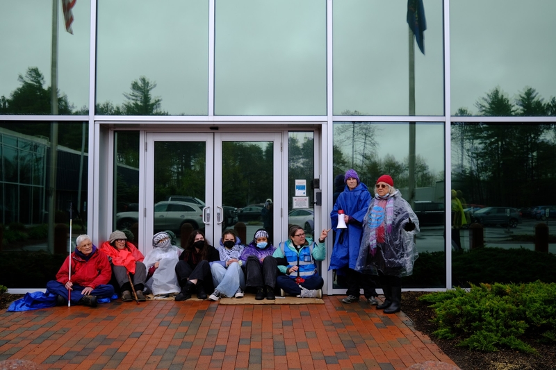 A group of people wearing rain gear sit and stand outside a modern glass building on a cloudy day.