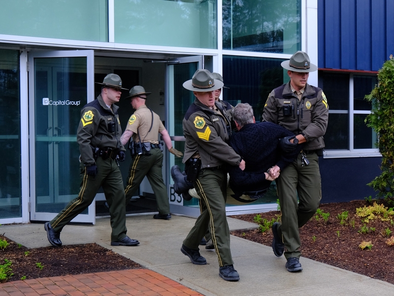 Four uniformed police officers carry a man out of a building through glass doors on a concrete walkway.
