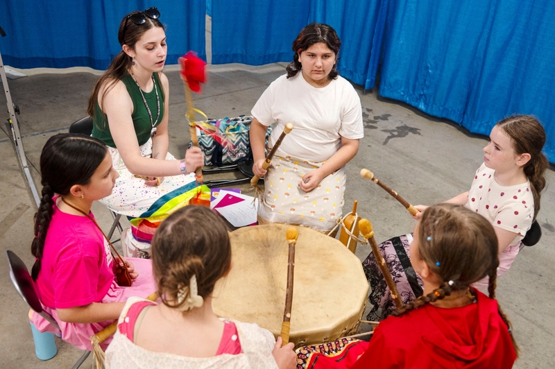 Six girls sit in a circle around a large drum, each holding a drumstick. One girl beats the drum with a red mallet. They are inside, near blue curtains.