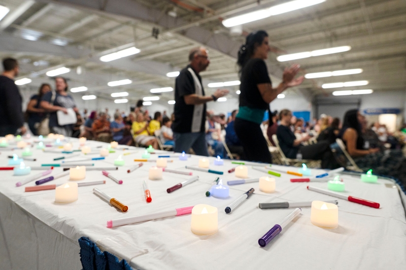 A table with electronic candles and marker pens in the foreground; people are seated and some are standing in a crowded indoor event space.