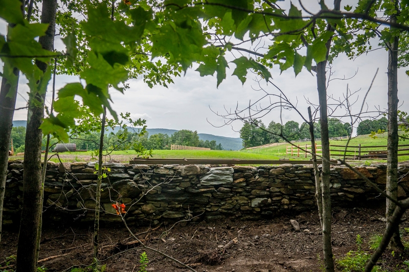 Stone wall with leafy trees in the foreground and green rolling hills, grassy fields, and fences visible in the background under a cloudy sky.