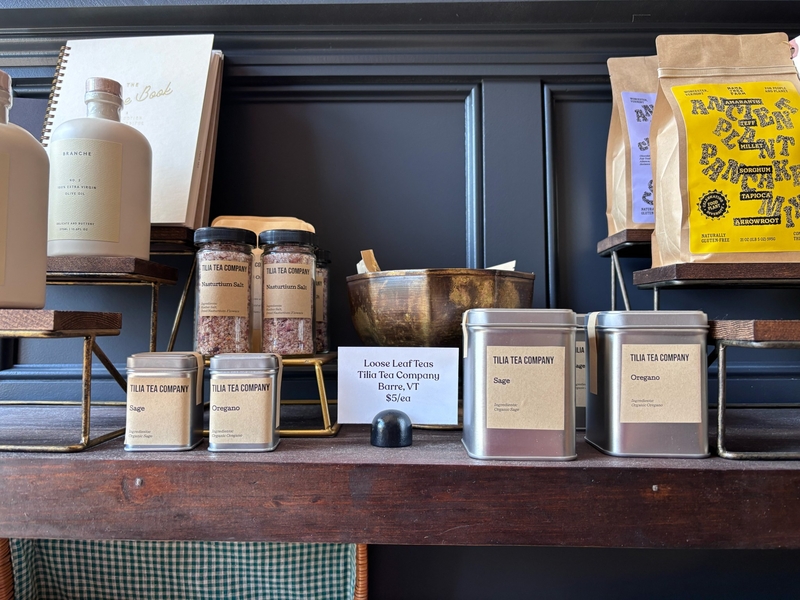 Jars, tins, and a bowl of tea and spice products displayed on a wooden shelf, with product labels and prices visible.