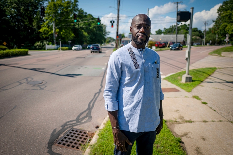 A man stands on a sidewalk near a busy intersection on a sunny day, wearing a light blue shirt with patterns on the collar and pocket. Trees and traffic lights are visible in the background.