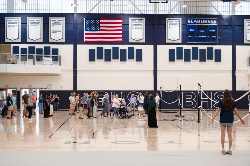 People gather in a high school gymnasium with a volleyball net set up, an American flag, and a scoreboard reading “Seahorses” visible on the wall.