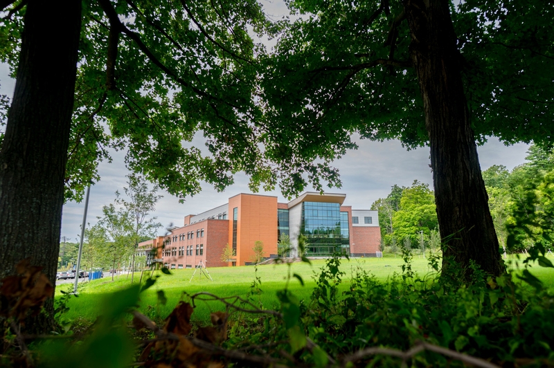 A modern brick and glass building is partially visible through large green trees, set on a grassy lawn under a cloudy sky.