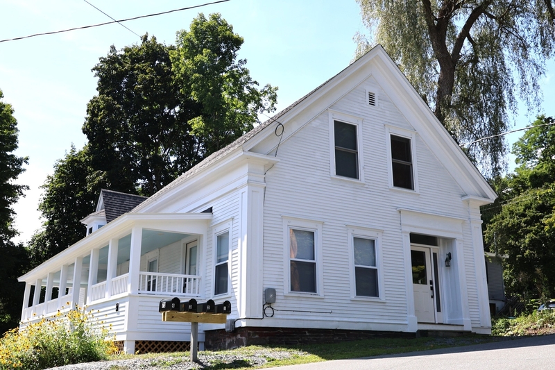 White two-story house with a covered wraparound porch, four black mailboxes in front, surrounded by trees and greenery on a sunny day.