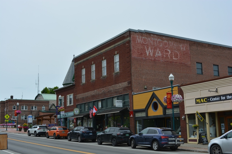 A row of brick and wood storefronts along a small town street, with parked cars and a faded “MONGOMERY WARD” sign on one building.
