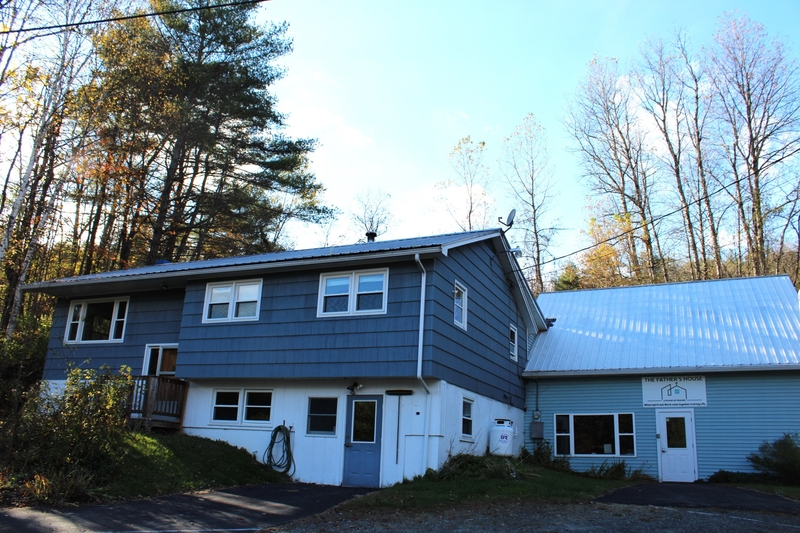 A blue two-story house with an attached building, white trim, a driveway, sparse autumn trees, and a clear sky in the background.