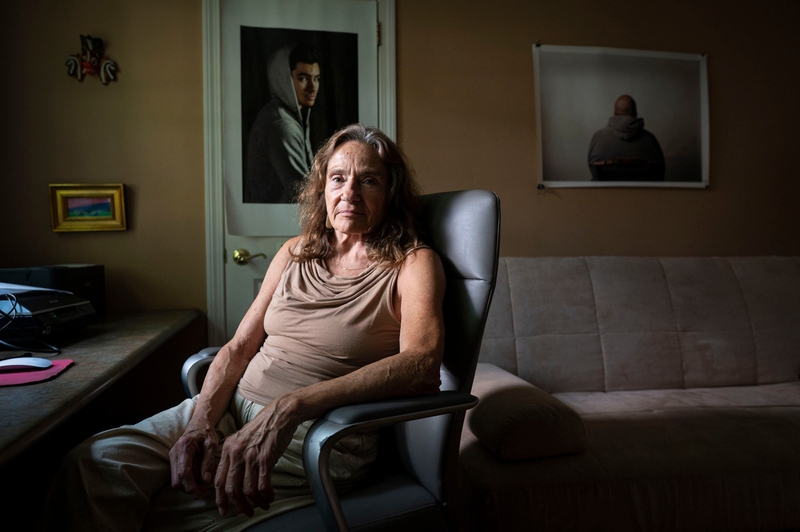 An older woman sits in an office chair in a dimly lit room with a couch, desk, and two portrait photographs on the walls behind her.
