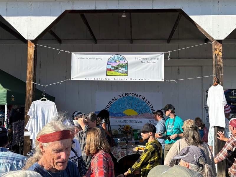 A group of people gather at an outdoor booth under a banner for Rural Vermont Rising, with shirts and informational materials displayed on tables.