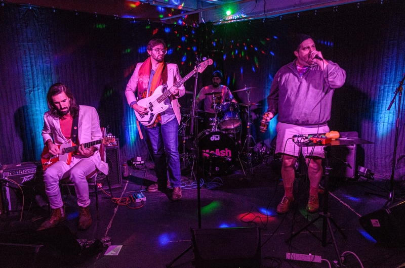 Four musicians perform on stage with guitar, bass, drums, and vocals under colorful lights and a dark backdrop.