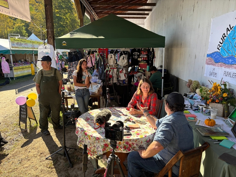 People sit at a table with recording equipment, while others stand nearby at an outdoor market stall displaying goods and informational signs.