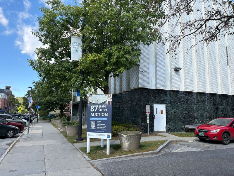 Sidewalk view with parked cars, trees, and a large sign announcing a property auction at 87 State Street in front of a modern office building with a marble and white exterior.
