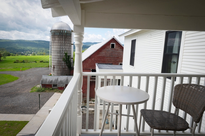 A small table and a wicker chair sit on a white porch overlooking a red barn, a silo, and green fields under a partly cloudy sky.