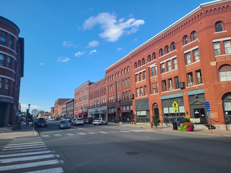 A city street lined with red brick buildings, parked cars, a crosswalk, and a clear blue sky above.