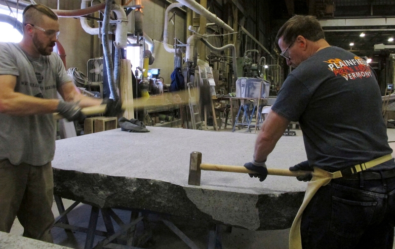 Two men use sledgehammers to split a large slab of stone in an industrial workshop, surrounded by machinery and tools.