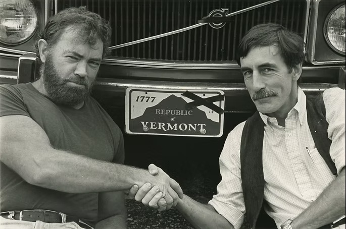 Two men shake hands in front of a car with a Vermont license plate altered to read “Republic of Vermont.”.