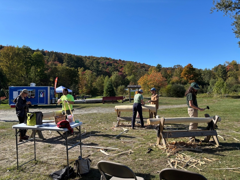People using hand tools to shape wooden logs at an outdoor workshop with a food truck in the background.
