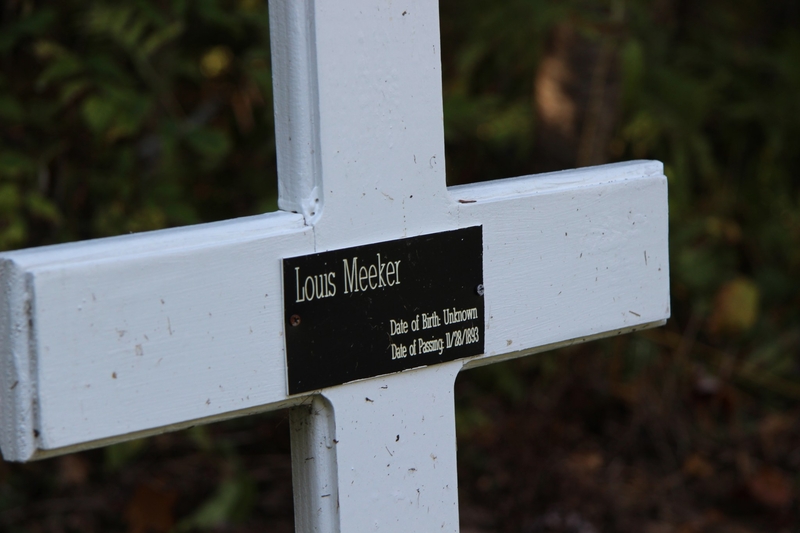 White wooden cross with a memorial plaque reading “Louis Meeker, Date of Birth: Unknown, Date of Passing: 11/28/1893.”