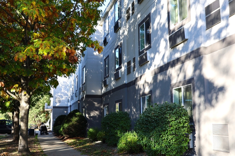 Side view of a multi-story hotel with window air conditioning units, adjacent to a sidewalk and a tree with autumn foliage.