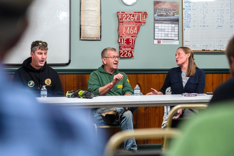 Three people sit at a table during a meeting, with the man in the center speaking and gesturing.