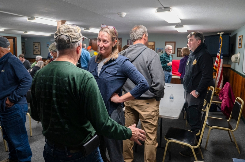 A man in a camouflage cap and green long-sleeve shirt talks to a woman in a blue button-down shirt at a community gathering.