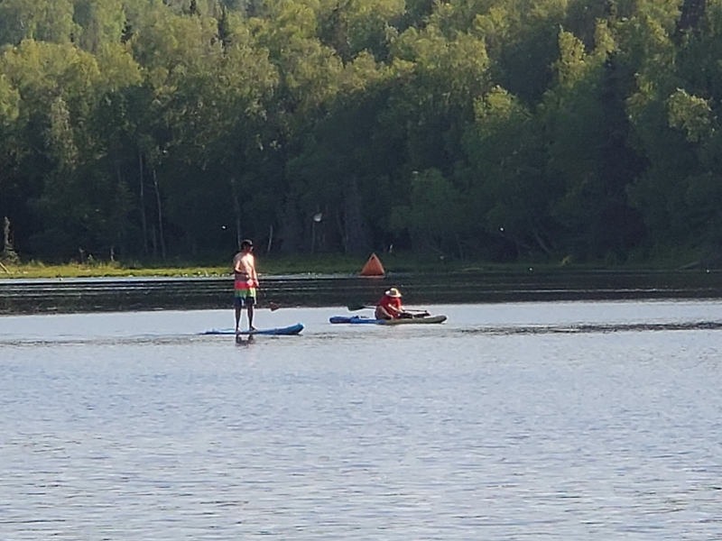 A paddleboarder and kayaker travel on Little Campbell Lake on June 22, 2022. The lake, within Kincaid Park, is a popular summer gathering site for swimmers, boaters, sunbathers and fishers. It is one of the sites in Alaska's largest city containing PFAS contaminants, according to a study by the nonprofit Alaska Community Action on Toxics. (Photo by Yereth Rosen/Alaska Beacon)