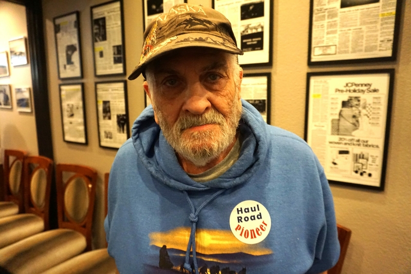 Fred Agree of Homer, wearing a sticker identifying himself as a "Haul Road Pioneer" who worked 50 years ago building the route now known as the Dalton Highway, stands by framed newspaper pages at the Petroleum Club of Anchorage on April 29, 2024. An exhibit marking the 50th anniversary of the Haul Road's consturction was set up at the club by the Alaska Oil and Gas Historical Society. (Photo by Yereth Rosen/Alaska Beacon)