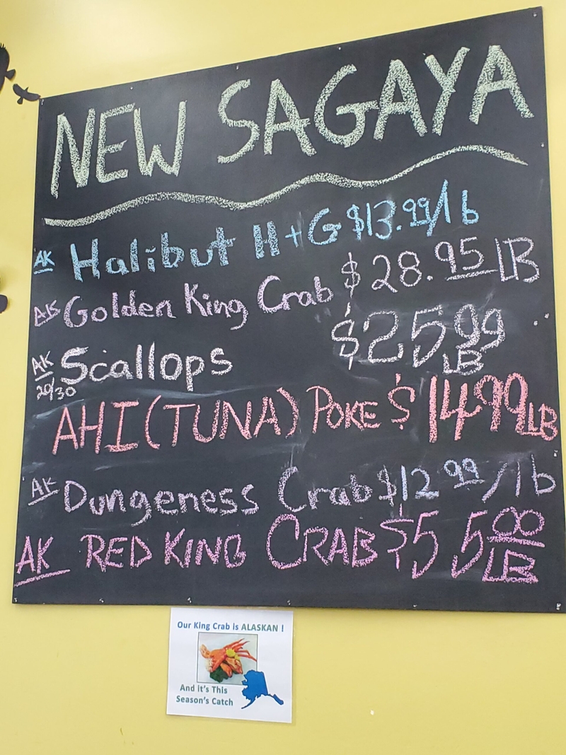 A blackboard at New Sagaga City Market in downtown Anchorage posts prices for crab and other fish on Dec. 1, 2023. A smaller sign celebrates the return of the Bering Sea red king crab harvest after a two-year closure in the Bristol Bay area. Red king crab, known for their huge sized, are iconic Alaska seafood that can fetch high prices. (Photo by Yereth Rosen/Alaska Beacon)