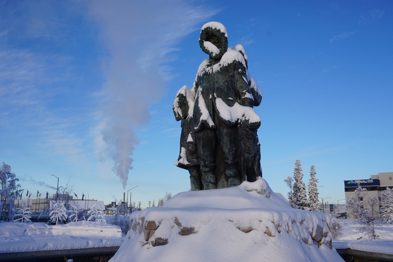 Smokestack emissions rise into the air on Feb. 7, 2025, above Golden Heart Plaza in downtown Fairbanks. Snow dusts the statute of the "Unknown First Family" that is the plaza's centerpiece. (Photo by Yereth Rosen/Alaska Beacon)