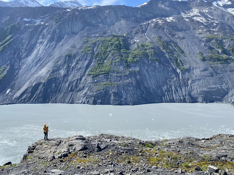 U.S. Geological Survey scientist Brian Collins evaluates a rock ledge on June 15. 2021, as a possible site to install equipment for monitoring movement of the Barry Arm landslide in Prince William Sound. The landslide is shown in the background across the fjord. (Photo by Dennis Staley/U.S. Geological Survey)