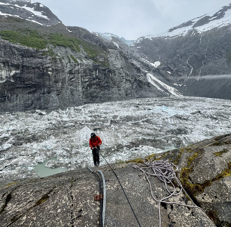 A U.S. Geological Survey scientist rappels down a rock face on ropes above Suicide Basin on June 27, 2025. The glacier-dammed lake is within the Mendenhall Glacier system above Juneau. Snow-covered mountains and the glacier are visible in the background, with coiled rope visible on the rocky ledge. (Photo provided by U.S. Geological Survey)