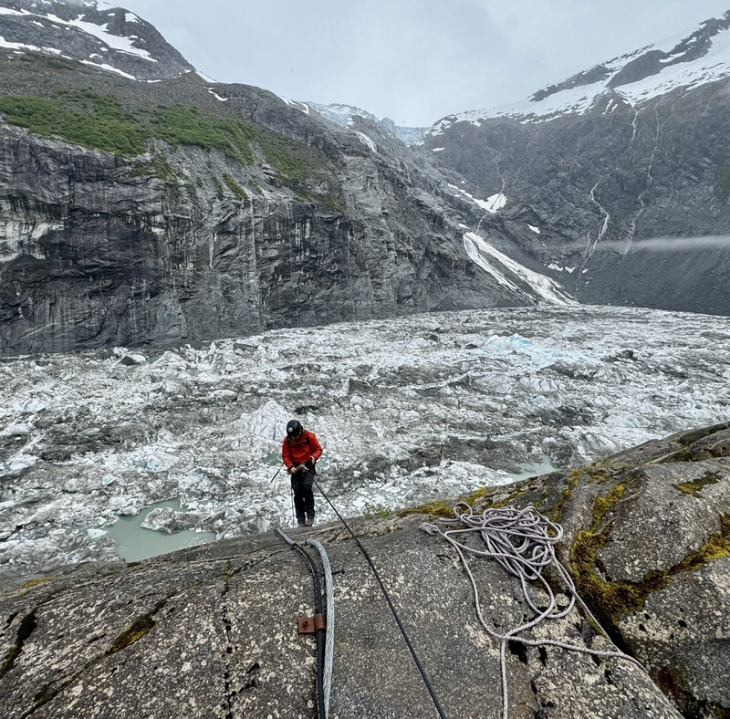 A U.S. Geological Survey scientist rappels down a rock face on ropes above Suicide Basin on June 27, 2025. The glacier-dammed lake is within the Mendenhall Glacier system above Juneau. Snow-covered mountains and the glacier are visible in the background, with coiled rope visible on the rocky ledge. (Photo provided by U.S. Geological Survey)