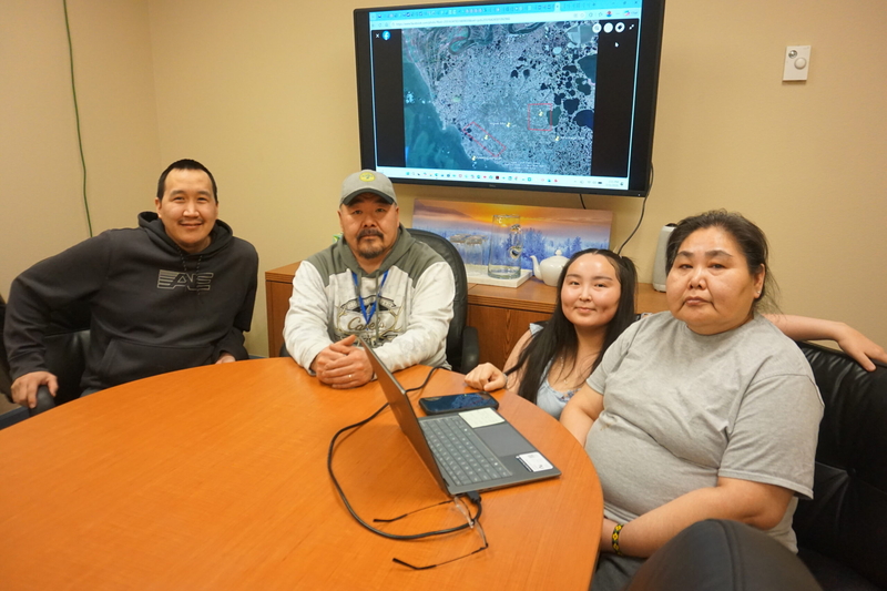 Rayna Paul and other Kipnuk tribal members sit at their temporary Anchorage office on Feb. 11, 2026. On the screne is a map of Kipnuk's current site and potential relocation sites. From left are Dolan Fox, Darren John, Carrie Dock and Paul. (Photo by Yereth Rosen/Alaska Beacon)