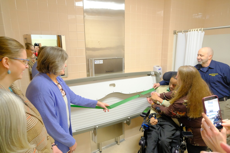 Judy and Eric Edwards gather with Reps. Sara Hannan, D-Juneau and Ashley Carrick, D-Fairbanks, and Capitol facilities manager Serge Lesh for a ribbon cutting ceremony for the installation of a new universal changing station at the Alaska State Capitol on Mar. 20, 2026. (Photo by Corinne Smith/Alaska Beacon)